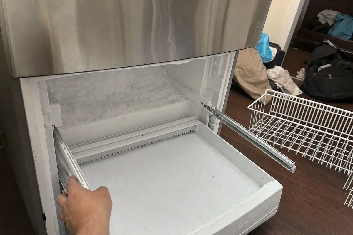 A hand drawing the frost-lined freezer drawer out of a stainless built-in, a wire basket lifted out onto the floor beside it
