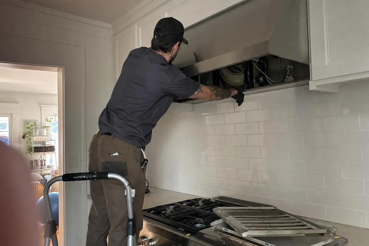 A technician on a step stool reaching up into the underside of a stainless hood, its filters out, the gas rangetop below him