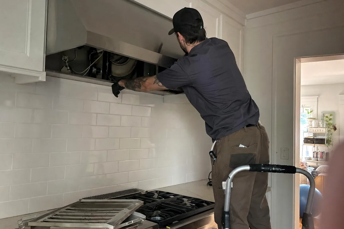 A stainless gas rangetop with its grates lifted off, seen from the side while a technician works at the hood above it