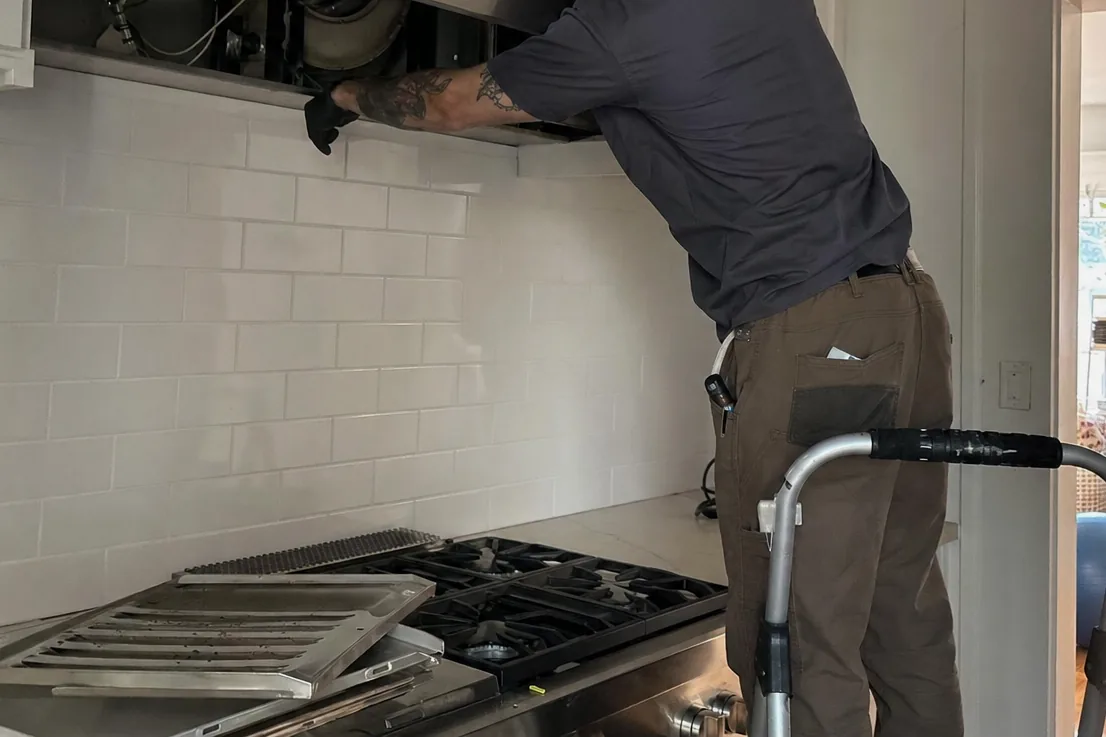 A service visit under way in a white tiled kitchen, the technician up at the hood, a doorway through to the next room behind him
