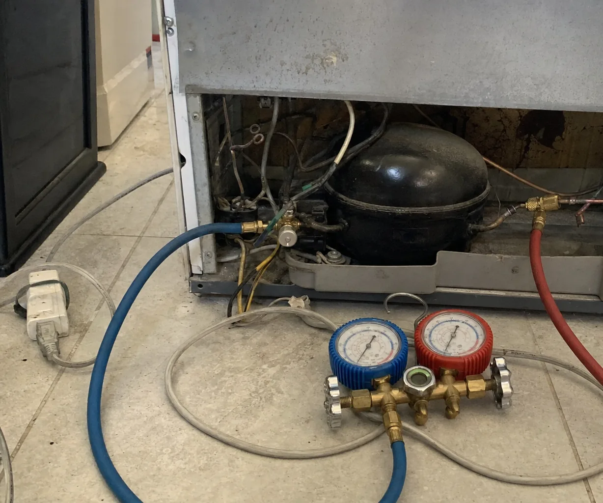 Refrigerator with its lower back panel removed, showing a black compressor and loose wiring, with blue and red gauge hoses running to a brass manifold on a tile floor