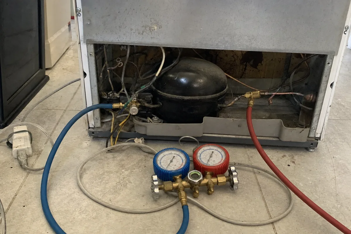 The open machine compartment of a built-in appliance with the compressor and copper lines exposed, blue and red manifold gauges connected on the floor in front of it