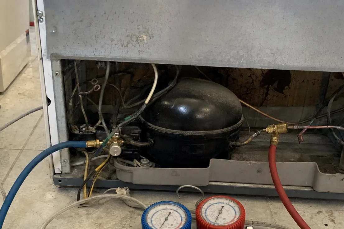 Manifold gauges with blue and red hoses lying on a tiled floor beside an opened appliance base
